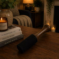 A dark charcoal-colored cast iron skillet with a wooden handle rests on a wooden table in a cozy, warmly lit room. The skillet is styled next to a stack of books and a lit candle in a ceramic jar.