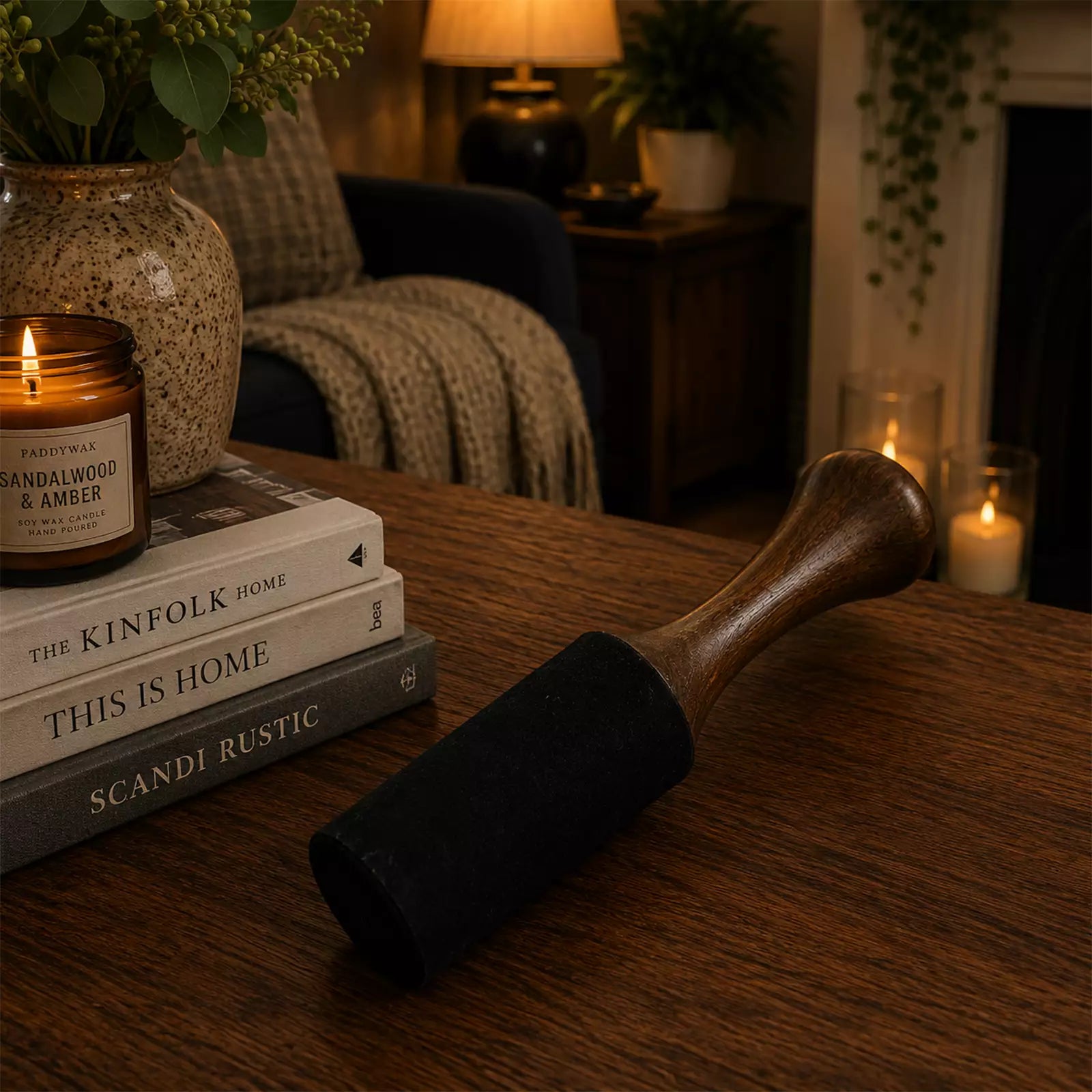 A dark charcoal-colored cast iron skillet with a wooden handle rests on a wooden table in a cozy, warmly lit room. The skillet is styled next to a stack of books and a lit candle in a ceramic jar.