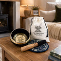 A singing bowl set on a wooden table in a cozy living room setting, featuring a black and gold-toned bowl, a wooden mallet, a blue patterned coaster, and a white drawstring bag printed with 'THE TAN LOTUS Singing Bowl'.