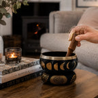 Hand holding wooden striker over Large Moon Phase Singing Bowl Set- Black 14cm with moon phase designs, placed on decorative cushion on a wooden table near lit candle and books in cozy living room setting.