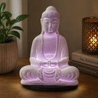 A seated Buddha figurine lamp glowing with soft purple light on a wooden table, styled with a potted plant on the left and a lit decorative candle on books to the right.