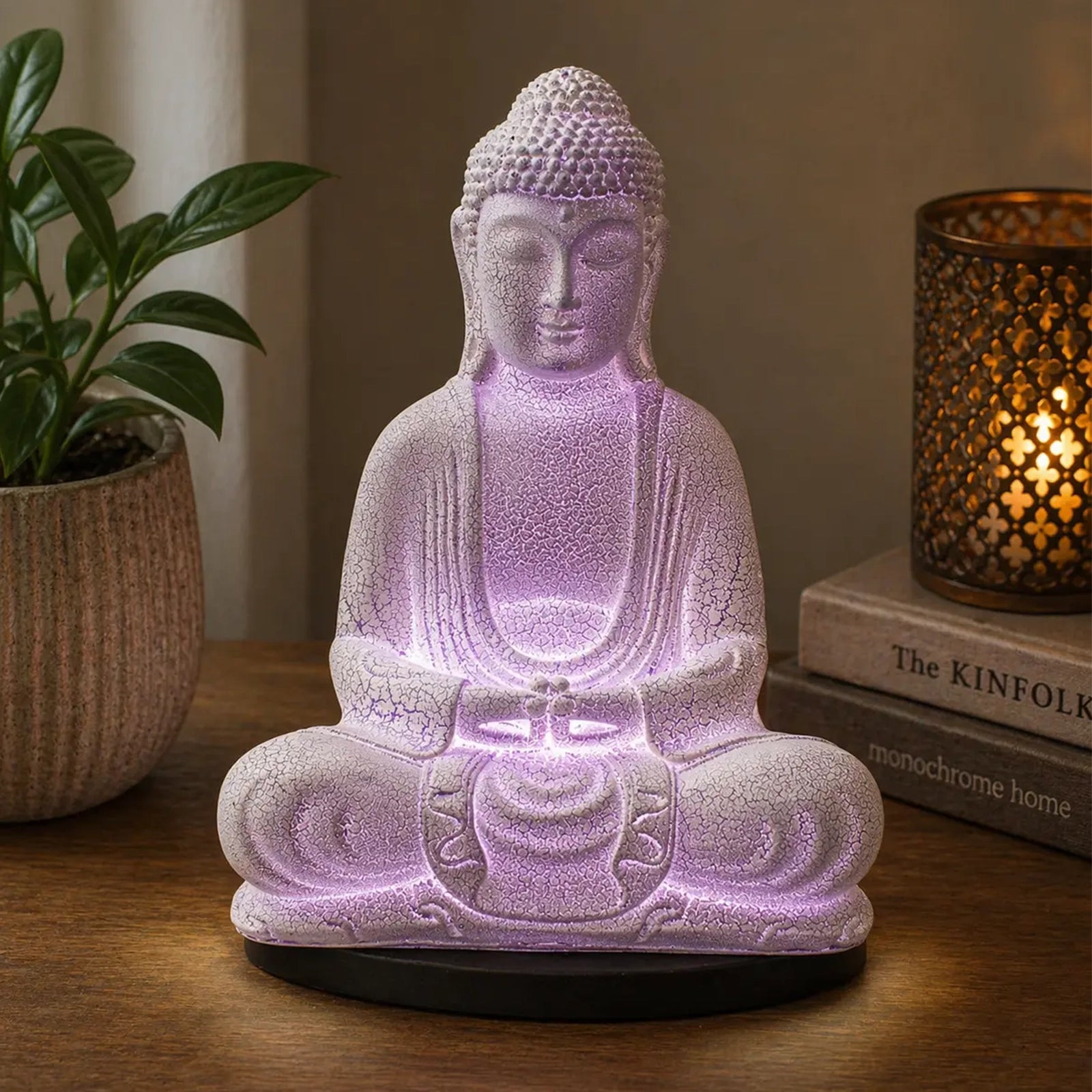 A seated Buddha figurine lamp glowing with soft purple light on a wooden table, styled with a potted plant on the left and a lit decorative candle on books to the right.