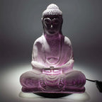 A purple glittery Buddha statue in a seated meditation pose, illuminated from within to cast a soft glow, shown against a dark gray background with its power cord visible on the right.