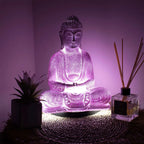 A glowing purple LED Buddha statue figurine in a seated meditation pose, styled on a woven mat with a small succulent and a reed diffuser bottle in a dimly lit room.