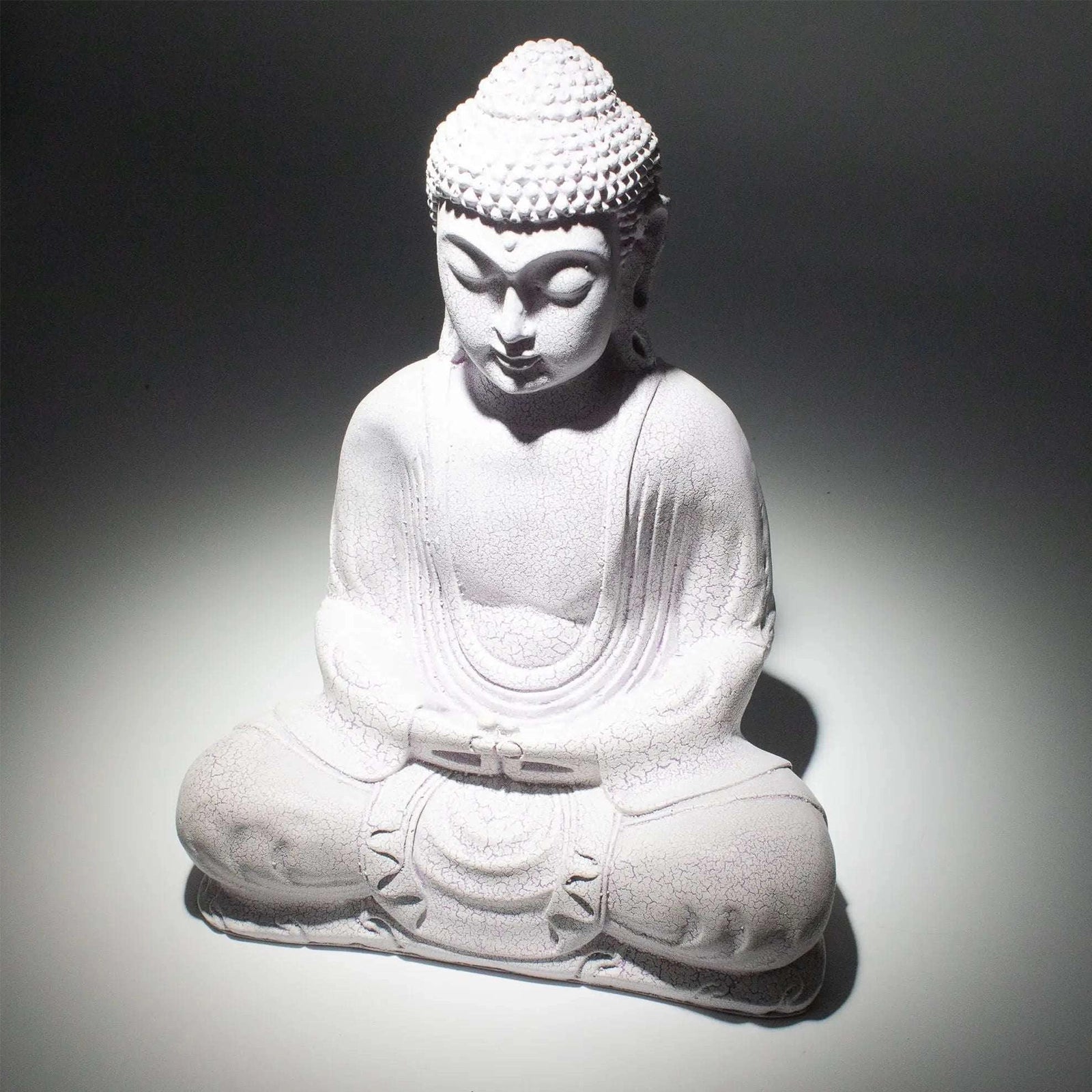 A white seated Buddha statue with a textured stone-like finish, photographed against a dark gray to white spotlight background.