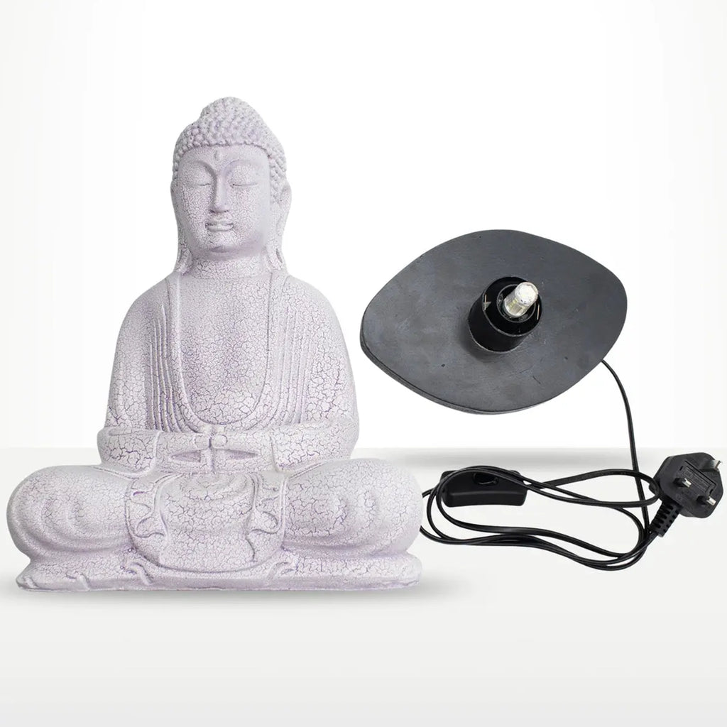 A white Buddha statue in a seated meditation pose shown next to its black circular light fixture with a power cord and UK plug, on a light gray background.