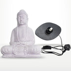 A white Buddha statue in a seated meditation pose shown next to its black circular light fixture with a power cord and UK plug, on a light gray background.