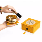 A brass singing bowl resting on a yellow cushion, with a hand holding a black-and-wood mallet above it. A matching yellow box with a decorative floral inlay and button closure sits to the right on a clean white background.