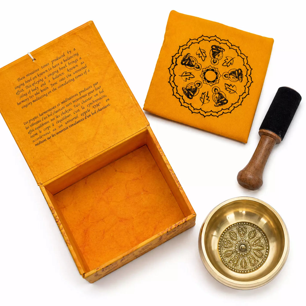 A brass singing bowl set with its open wooden box, a folded yellow fabric cover printed with a black mandala, a wooden mallet with a black felt head, and a small brass spoon on a white background.