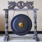 A large round gong with a gold-colored center, suspended from a dark gray carved wooden frame with dragon-like carvings at the top, set against a plain beige wall and tiled floor.
