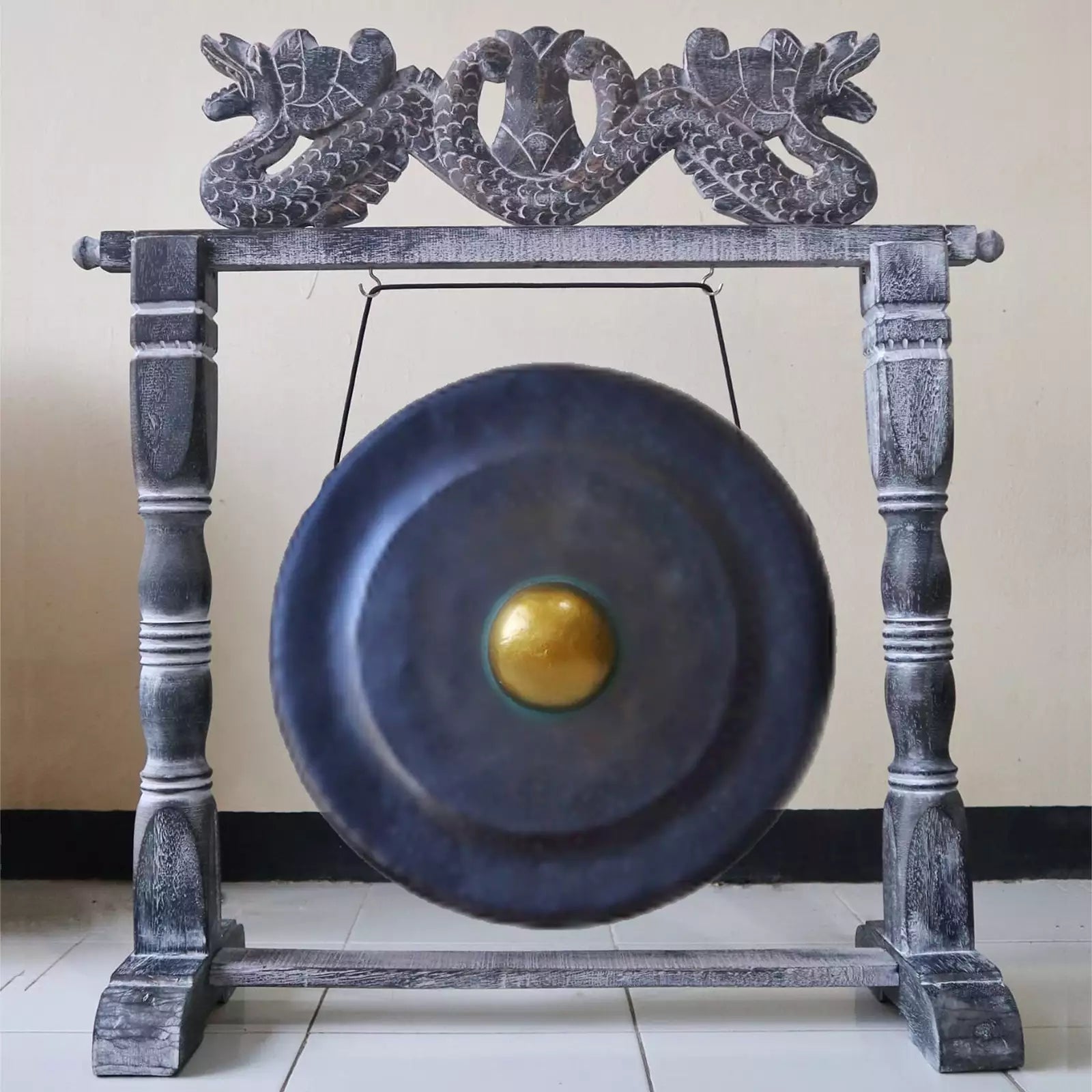 A large round gong with a gold-colored center, suspended from a dark gray carved wooden frame with dragon-like carvings at the top, set against a plain beige wall and tiled floor.