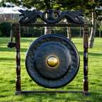 A large round bronze gong with a polished golden center, mounted on a dark wooden frame with dragon carvings at the top, set outdoors on a green lawn.