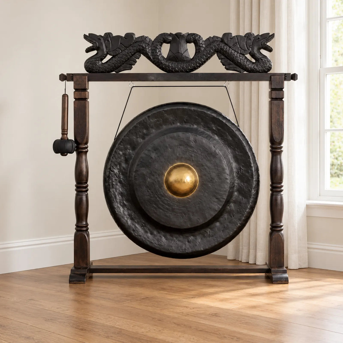 A large black metal gong with a gold-colored central boss, mounted on a dark wooden stand with dragon-shaped top carvings, shown in a bright room with wood flooring and a window.