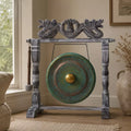 A decorative gong with a distressed gray metal frame and a round green patinated drum head, displayed indoors near a window with a beige chair and vase in the room.