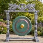 A large round bronze gong with a green patina and gold center, mounted on a weathered gray stone frame with dragon carvings, photographed outdoors in a garden setting.