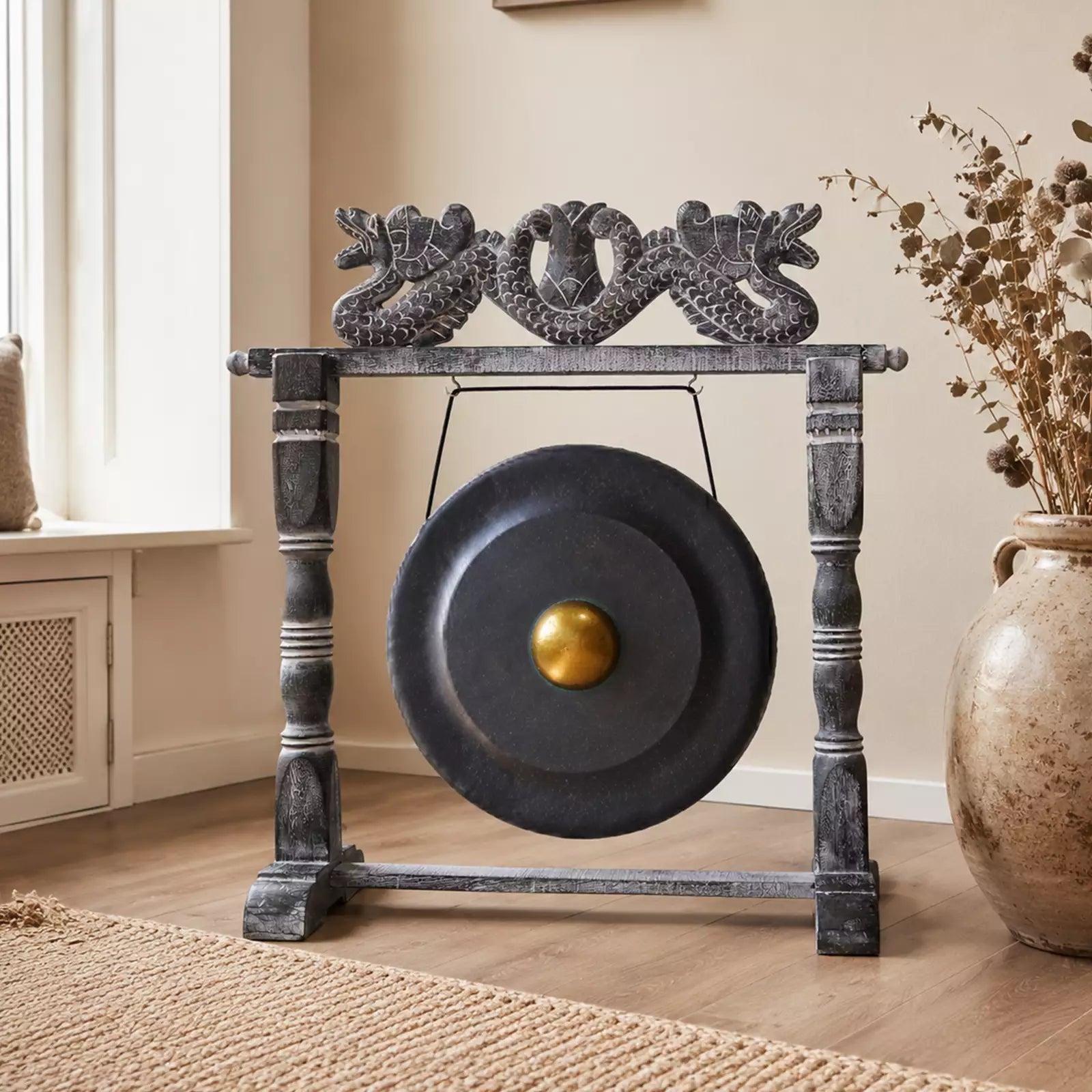 A large round black gong with a gold center, mounted on a dark gray decorative stand with dragon carvings at the top, shown in a styled room setting with a window, wooden floor, woven rug, and dried flowers.