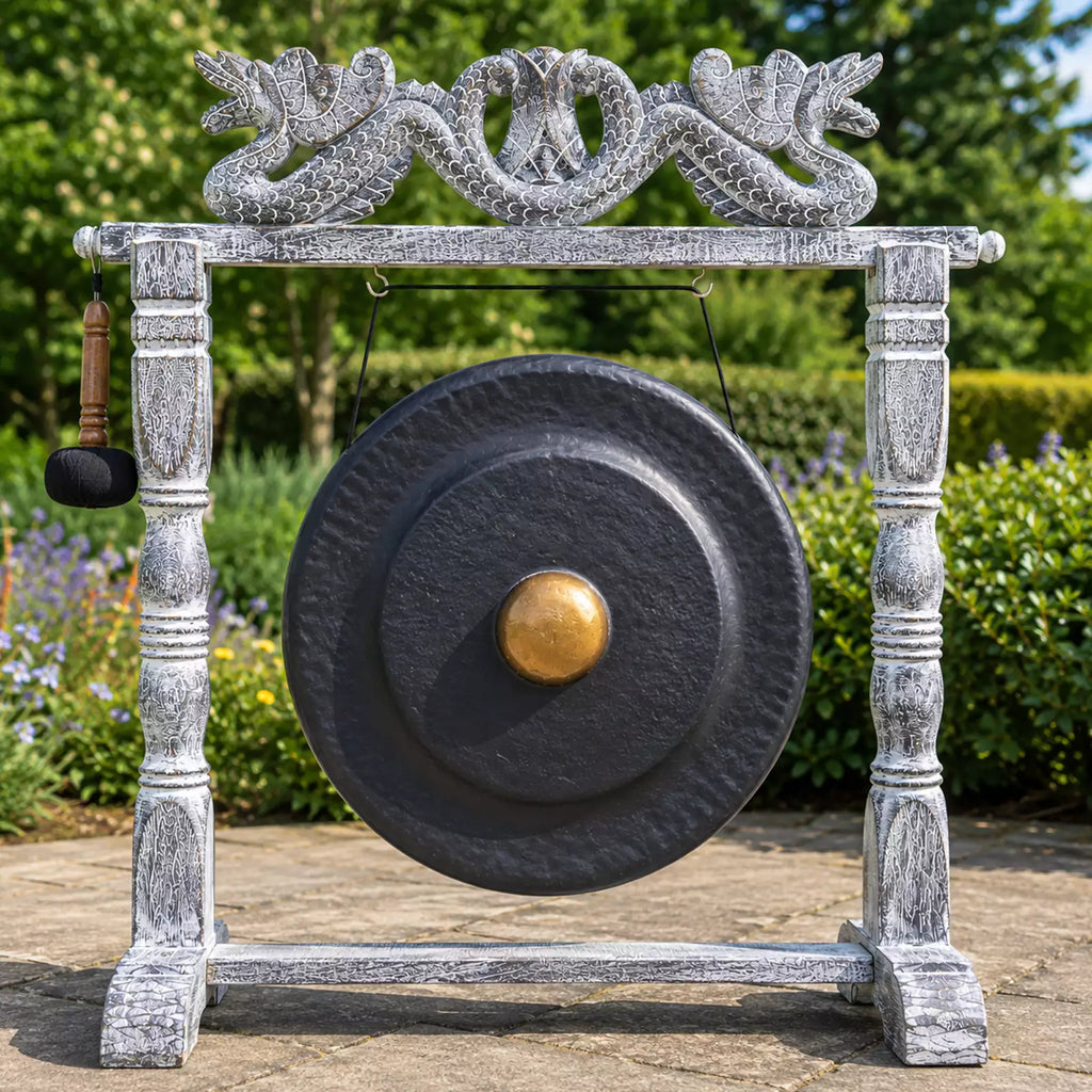 A large round black gong with a gold-colored center boss hangs from a distressed white wooden frame, with a hanging mallet on the left. The setup is photographed outdoors in a garden setting with green foliage and a stone path.
