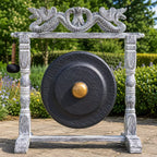 A large round black gong with a gold-colored center boss hangs from a distressed white wooden frame, with a hanging mallet on the left. The setup is photographed outdoors in a garden setting with green foliage and a stone path.