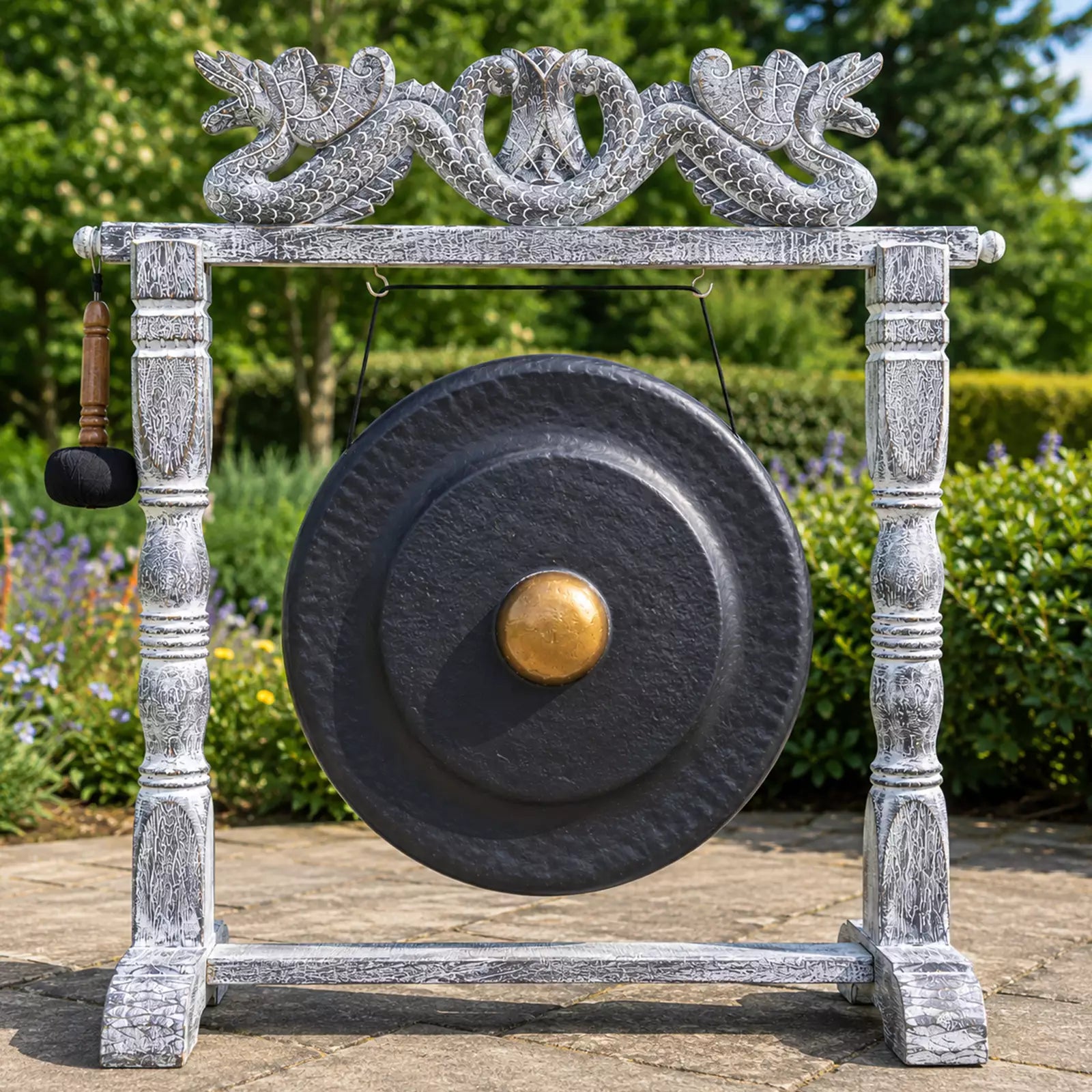 A large round black gong with a gold-colored center boss hangs from a distressed white wooden frame, with a hanging mallet on the left. The setup is photographed outdoors in a garden setting with green foliage and a stone path.