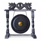A large round black gong with a gold-colored central boss, suspended from a dark wooden stand featuring ornate dragon carvings at the top, isolated on a white background.