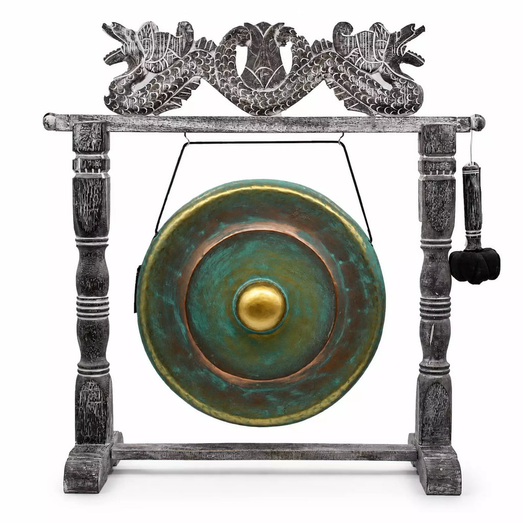 A decorative gong set on a carved stand, featuring a round green and bronze gong with a gold center, suspended by a wire from a weathered gray wooden frame topped with a dragon motif, and a hanging mallet on the right.
