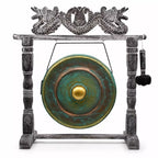A decorative gong set on a carved stand, featuring a round green and bronze gong with a gold center, suspended by a wire from a weathered gray wooden frame topped with a dragon motif, and a hanging mallet on the right.