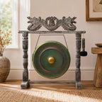 A decorative bronze gong with a distressed green patina and a gold center, mounted on a dark gray carved wooden stand with dragon details, set in a sunlit room on a woven rug.