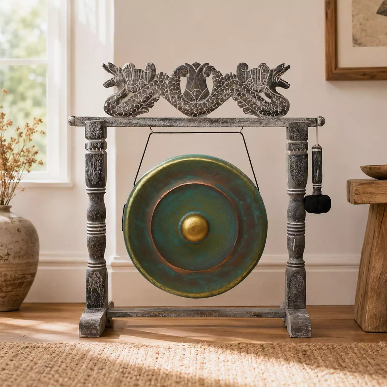 A decorative bronze gong with a distressed green patina and a gold center, mounted on a dark gray carved wooden stand with dragon details, set in a sunlit room on a woven rug.