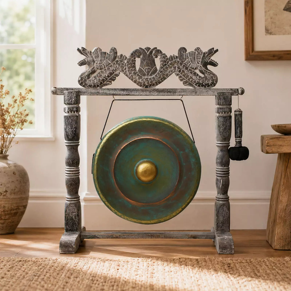 A decorative bronze gong with a distressed green patina and a gold center, mounted on a dark gray carved wooden stand with dragon details, set in a sunlit room on a woven rug.