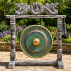 A large round gong with a turquoise-green and bronze patina finish hangs from a weathered gray stone-style frame decorated with dragon sculptures at the top. A black hanging mallet is attached to the right side.