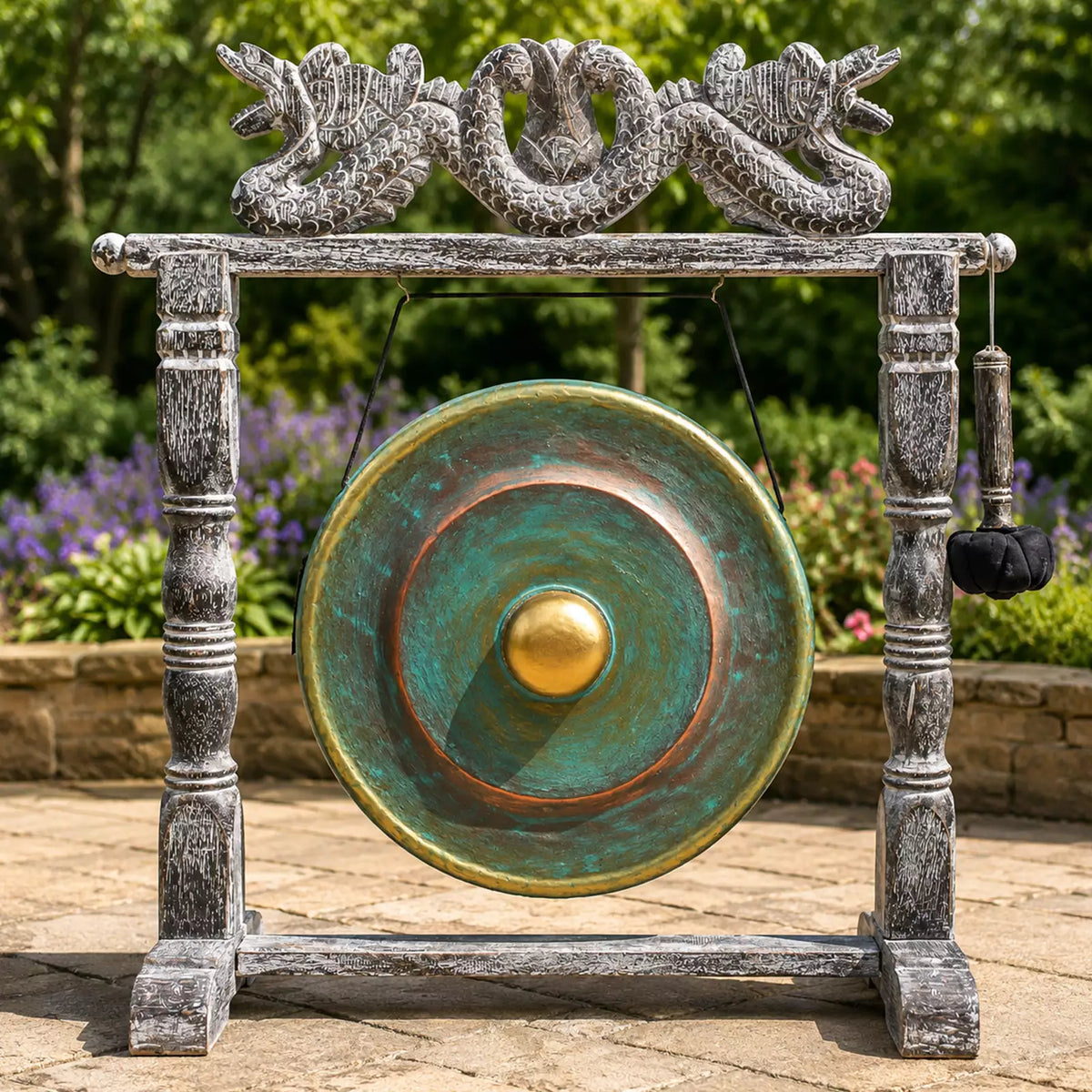 A large round gong with a turquoise-green and bronze patina finish hangs from a weathered gray stone-style frame decorated with dragon sculptures at the top. A black hanging mallet is attached to the right side.
