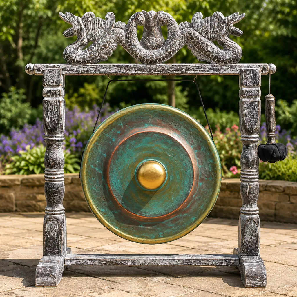 A large round gong with a turquoise-green and bronze patina finish hangs from a weathered gray stone-style frame decorated with dragon sculptures at the top. A black hanging mallet is attached to the right side.