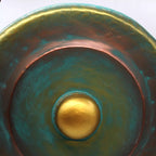 Close-up of a round ceramic bowl or planter with a textured metallic finish, featuring concentric rings of teal, copper-brown, and a central gold accent.