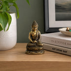 Mini Meditating Sitting Buddha in antique brass finish displayed on a wooden surface next to books and a white textured planter with a green leafy plant.