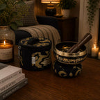 A decorative singing bowl set on a wooden table in a warmly lit living room. The set includes a round metal bowl with a dark wooden striker resting inside, both covered in black fabric with ornate gold patterns.