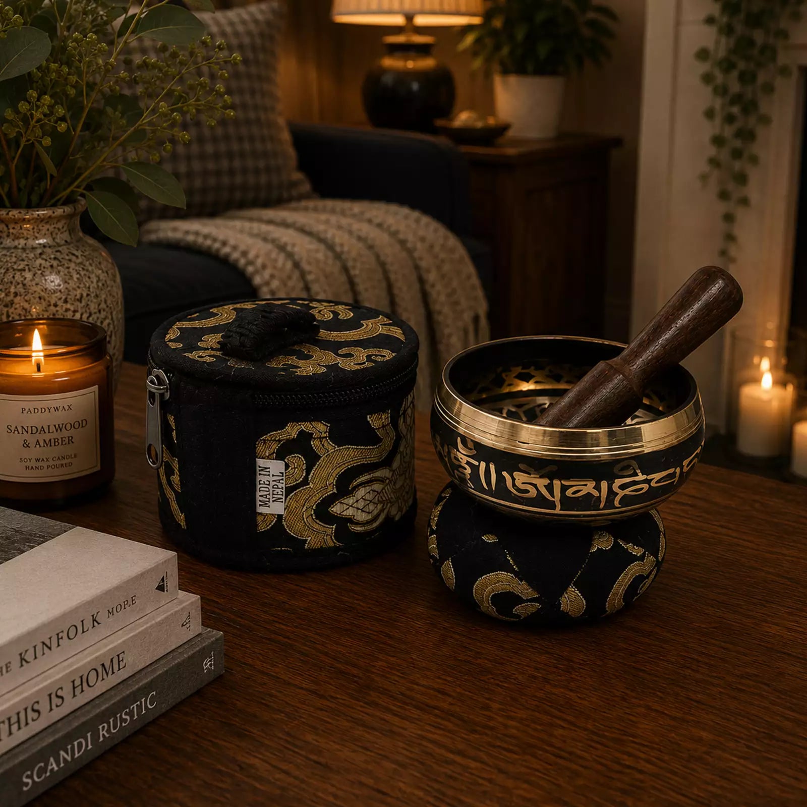 A decorative singing bowl set on a wooden table in a warmly lit living room. The set includes a round metal bowl with a dark wooden striker resting inside, both covered in black fabric with ornate gold patterns.
