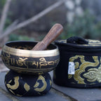 A brass singing bowl with gold Tibetan script, resting on a matching black and gold fabric stand with a wooden mallet inside, next to its black zippered carrying case with gold embroidery.