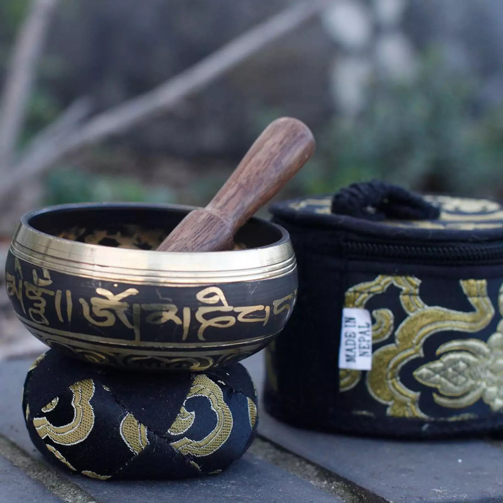 A brass singing bowl with gold Tibetan script, resting on a matching black and gold fabric stand with a wooden mallet inside, next to its black zippered carrying case with gold embroidery.