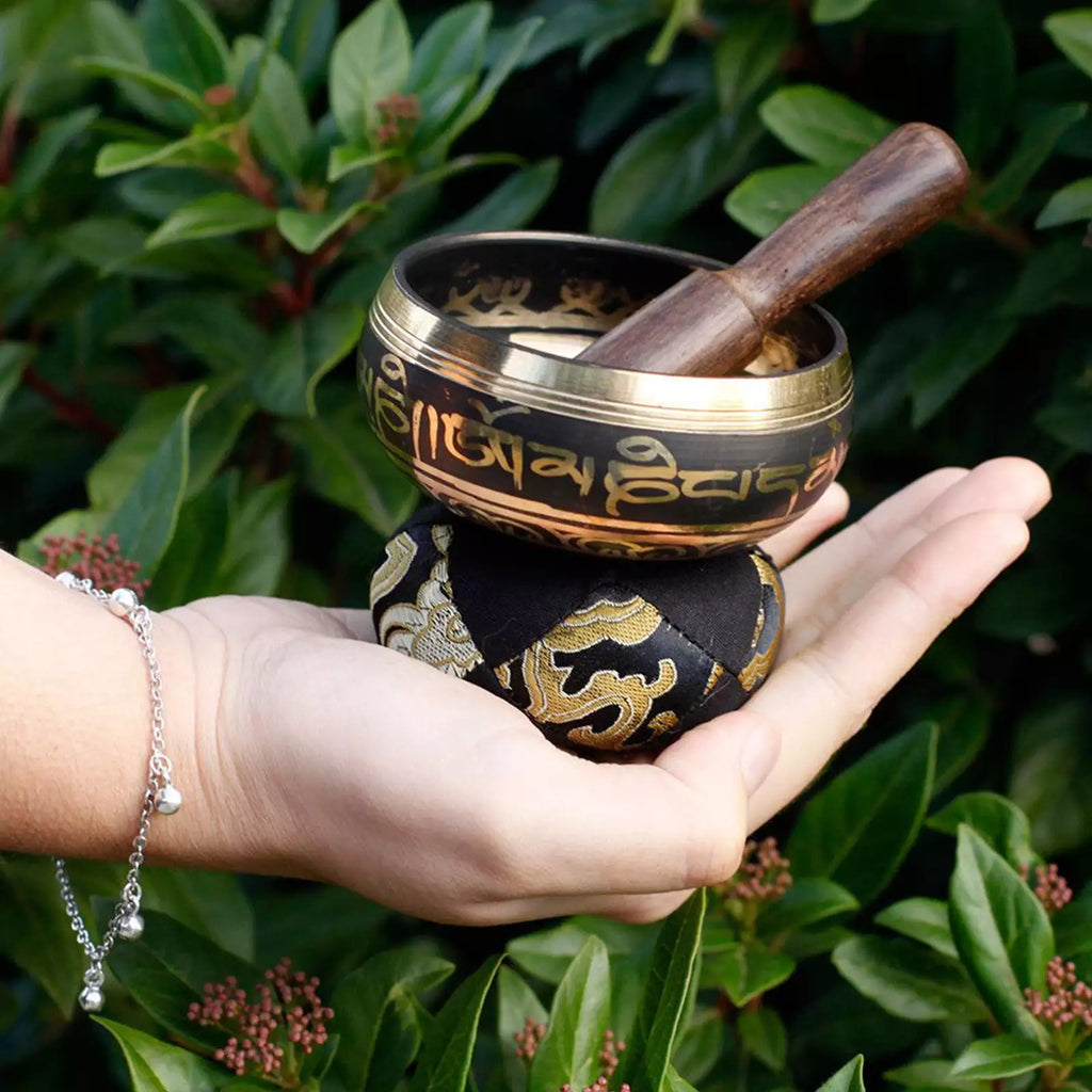 A Tibetan singing bowl set with a wooden mallet, resting on a black and gold patterned cushion held in a person's hand, with a background of green foliage.