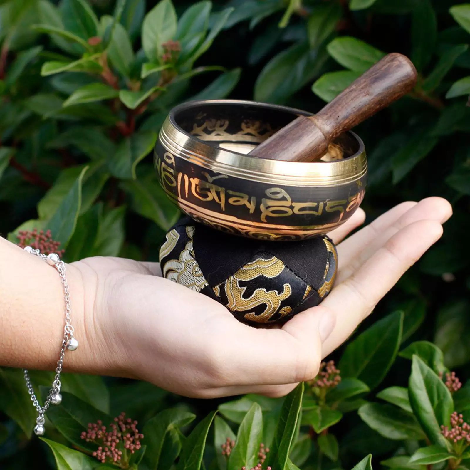 A Tibetan singing bowl set with a wooden mallet, resting on a black and gold patterned cushion held in a person's hand, with a background of green foliage.