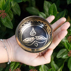 A hand holding a round metallic bowl with a black center and silver-toned decorative motifs, set against a background of green leaves and small red berries.