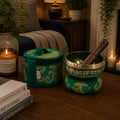 A green and gold Tibetan singing bowl set with a wooden mallet on a wooden table, styled in a cozy home environment with books, a candle, and plants.