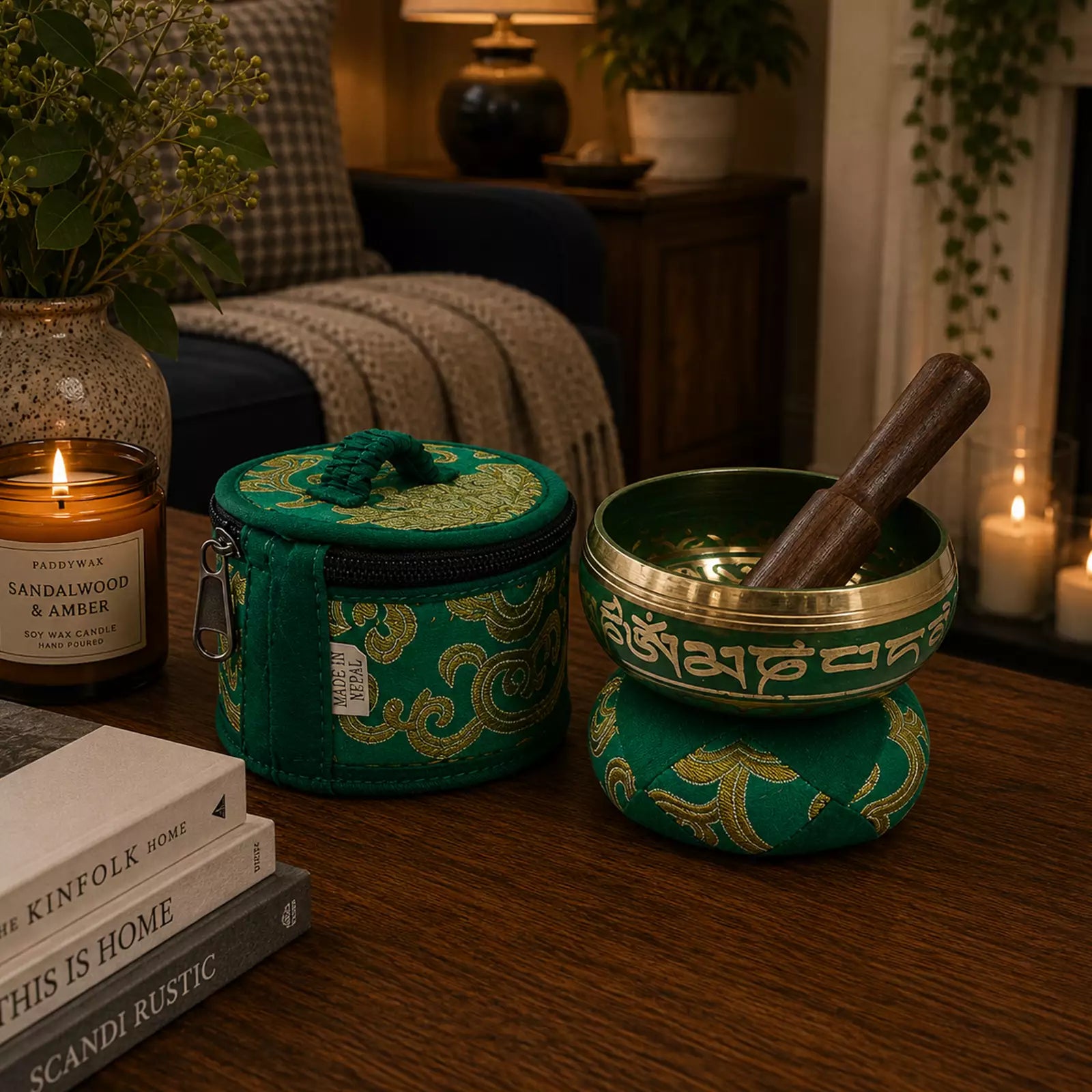 A green and gold Tibetan singing bowl set with a wooden mallet on a wooden table, styled in a cozy home environment with books, a candle, and plants.