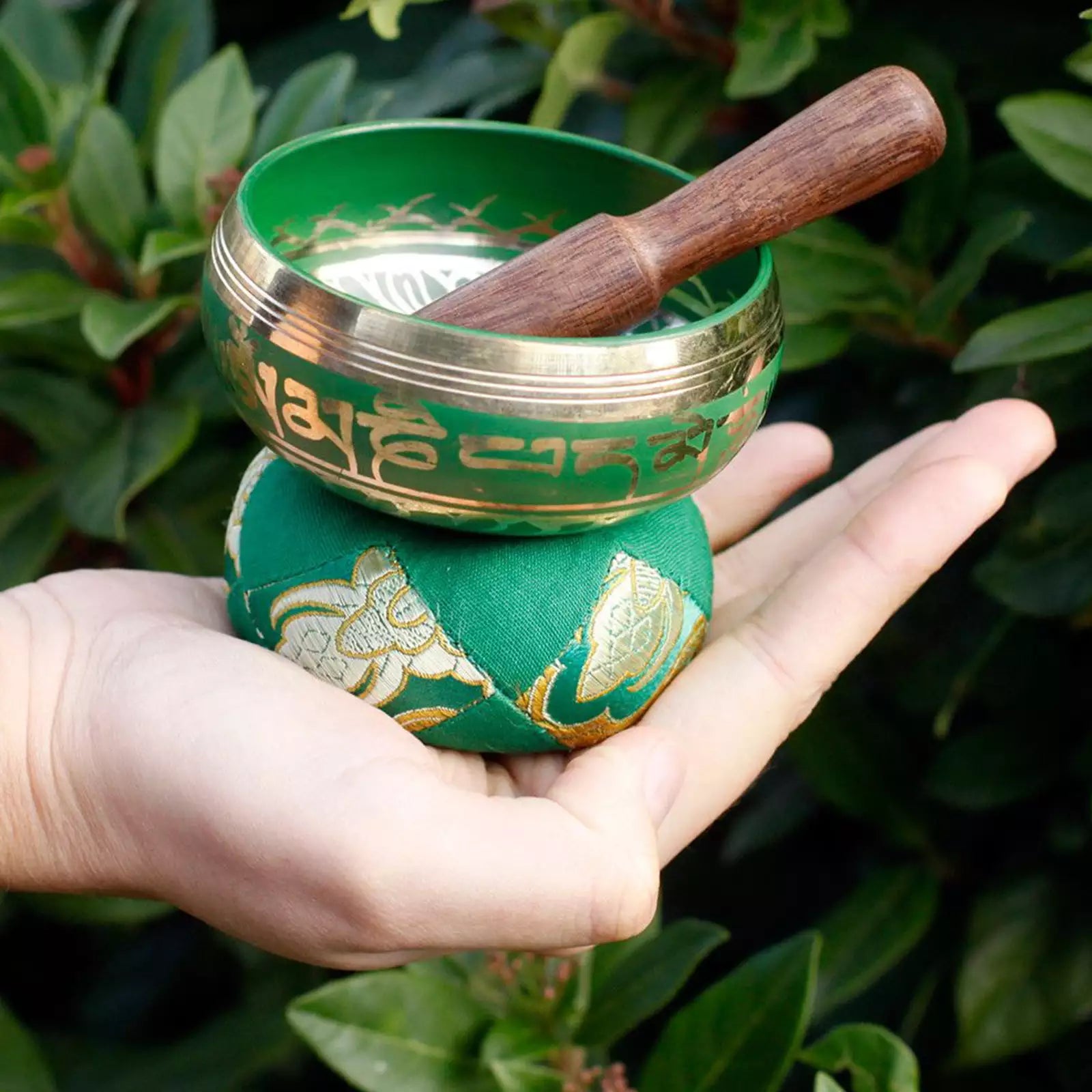 A green Tibetan singing bowl with gold-colored engraved script and a wooden mallet, held in a person's hand against a background of green foliage.
