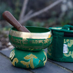 A green Tibetan singing bowl with gold script and a wooden mallet, resting on a matching green cushion, with a second cushion blurred in the background on an outdoor stone surface.