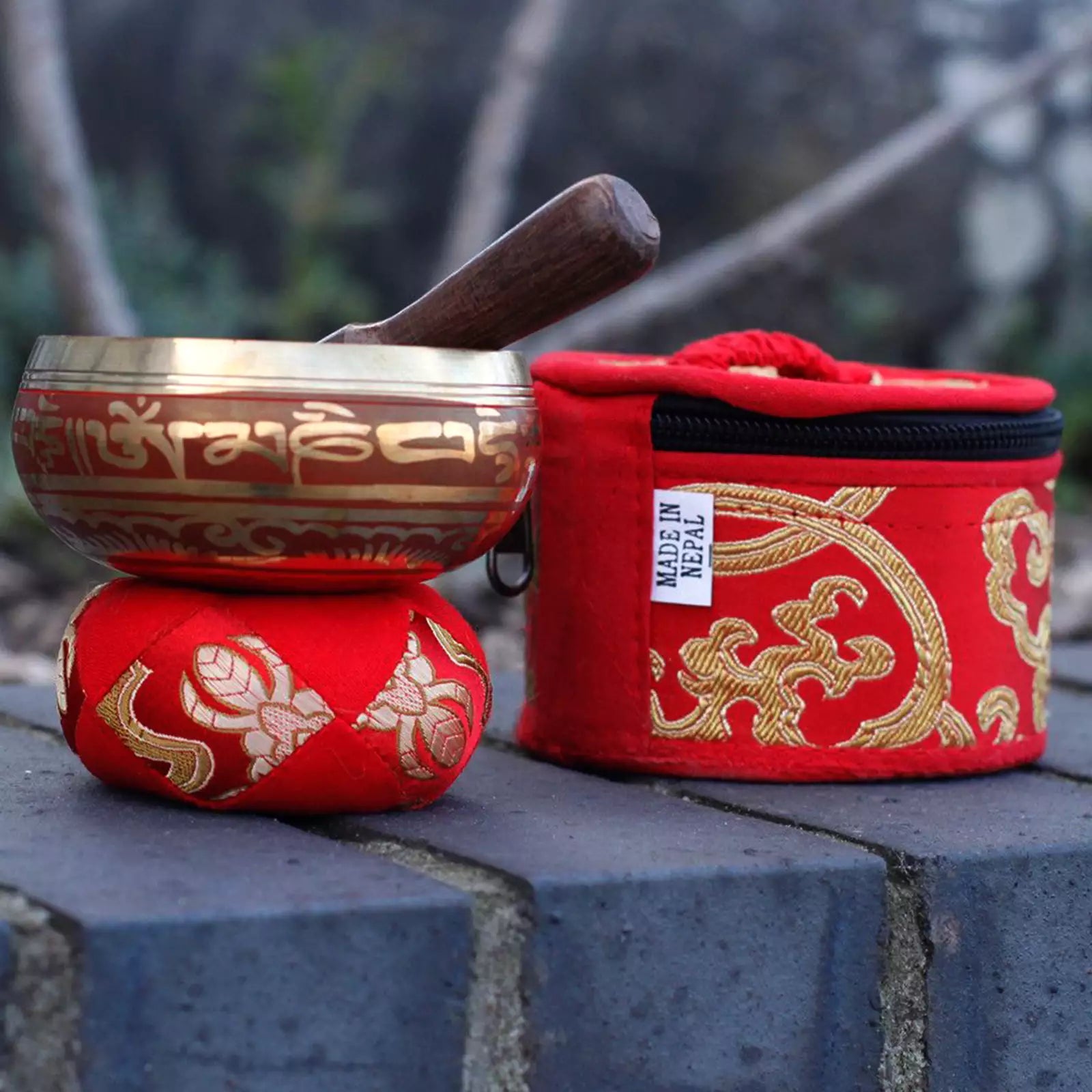 A Tibetan singing bowl set with a metallic bowl and wooden mallet resting on a red cushion, alongside a matching red zippered carrying case with gold embroidery and a visible 'MADE IN NEPAL' label, placed on a stone surface with a blurred outdoor background.