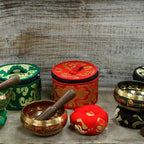 A set of Tibetan singing bowls and their matching fabric storage cases, featuring red, green, brown, and black colors with gold embroidered patterns, arranged on a rustic wooden surface.