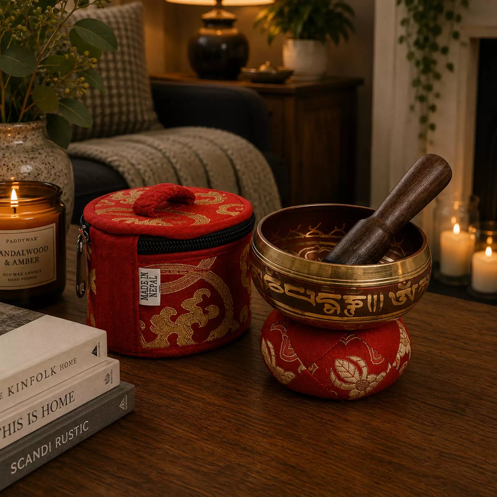 A brass singing bowl set with a wooden mallet resting inside, placed on a red patterned cushion on a wooden table. A matching red zippered storage case sits beside it, with candles, books, and home decor softly blurred in the background.