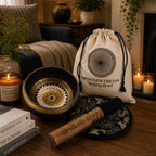 A singing bowl set displayed on a wooden table in a cozy room setting. The set includes a black Tibetan singing bowl with a gold geometric pattern inside, a wooden mallet, a black and gold patterned cloth coaster, and a cream-colored drawstring pouch labeled 'MEDALLION TIBETAN Singing Bowl'.