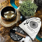 A singing bowl set arranged on a rustic wooden surface, including a black metal singing bowl with a gold-toned engraved interior, a white drawstring pouch printed with 'MEDULLION THERAPY Singing Bowl,' a black-and-white patterned cloth, and a wooden-handled striker. A potted green plant and decorative stones are also present.